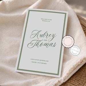 May include: A rectangular wedding invitation card with a light gray background and a dark green border. The text reads: WELCOME TO THE WEDDING OF Aubrey AND Thomas GOLD COAST, QUEENSLAND Saturday, 3rd of September. Two small circular stickers are attached.