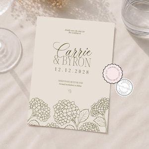 May include: A cream-colored wedding save the date card featuring olive green text that reads Carrie & BYRON 12.12.2028, Gold Coast, Queensland, and a line drawing of hydrangea flowers at the bottom, with two small circular stickers.