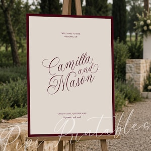 Op de afbeelding: Een rechthoekig welkomstbord voor een bruiloft met een crèmekleurige achtergrond en een donkerrode rand. De tekst luidt: WELCOME TO THE WEDDING OF, Camilla and Mason, GOLD COAST, QUEENSLAND, September 3rd, 2028. Het bord staat op een houten ezel.