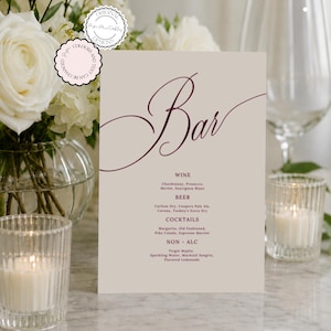 Könnte beinhalten: Eine beige Bar-Speisekarte mit burgunderfarbener Schrift, die oben "Bar" lautet. Die Speisekarte listet Wein, Bier, Cocktails und alkoholfreie Getränke auf. Die Speisekarte ist vor dem Hintergrund weißer Blumen, Kerzen und Glaswaren.