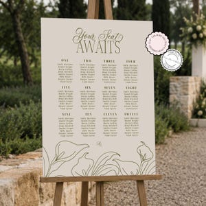 Peut inclure: Un panneau de plan de table de mariage beige sur un chevalet en bois avec le texte Your Seat AWAITS, une liste de douze tables avec des noms et une illustration au trait de lys calla en bas. Deux badges circulaires sont visibles dans le coin.