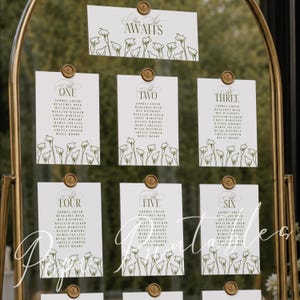 Könnte beinhalten: Ein goldener Bogenrahmen mit weißen Sitzplan-Karten, die mit grünen Calla-Lilien-Illustrationen verziert sind. Die Karten zeigen den Text Your Seat AWAITS und die Tischnummern eins bis sechs mit Namen in schwarzer Schrift, befestigt mit goldenen Clips.