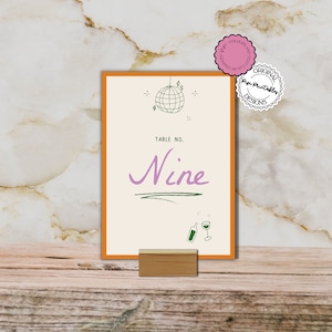Puede incluir: Tarjeta de número de mesa naranja y crema con una bola de discoteca verde, estrellas y copas de vino. La tarjeta dice "Table No. Nine" en texto verde y morado.