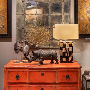 Könnte beinhalten: Eine rote lackierte Kommode mit drei Schubladen und schwarzen Metallgriffen. Auf der Kommode stehen eine schwarze Nashornfigur, drei afrikanische Masken, ein goldener Zweig und eine Lampe mit weißem Schirm und einem schwarz-weiß karierten Sockel.