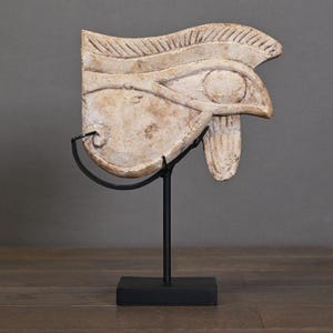 Könnte beinhalten: Beigefarbene altägyptische Horusauge-Skulptur. Die Skulptur ist auf einem schwarzen Metallständer mit rechteckigem Sockel montiert. Das Auge ist mit geschnitzten Linien und einer kreisförmigen Pupille detailliert.
