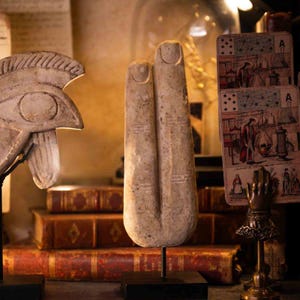 Könnte beinhalten: Eine beige Steinskulptur von zwei Fingern, montiert auf einem schwarzen Ständer. Die Skulptur hat geätzte Linien und ist vor dem Hintergrund antiker Bücher und Spielkarten platziert. Das Gesamtthema ist antike Kunst.