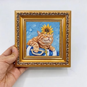 May include: An oil painting in a decorative gold frame depicting an orange tabby cat with a yellow sunflower on its head. The cat rests its paws on a blue and white striped object against a textured light blue background with visible brushstrokes.
