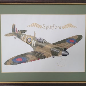 May include: Cross-stitch artwork of a Spitfire airplane in flight, framed in a dark brown frame with a green mat. The plane is detailed with camouflage, roundels, and the word "Spitfire" above it.