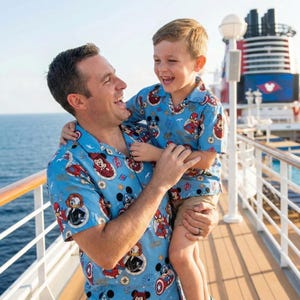 Op de afbeelding: Bijpassende lichtblauwe overhemden met een Disney- en Marvel-personageprint. De overhemden worden gedragen door een persoon en een kind, beiden glimlachend. De achtergrond toont een cruiseschip en de zee.