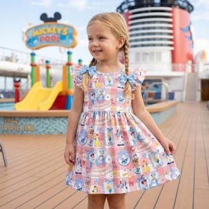 Op de afbeelding: Een jong meisje draagt een roze en witte jurk met een patroon en blauwe strikken in haar haar. De jurk heeft illustraties van Disney-personages. De achtergrond toont een dek van een cruiseschip en een zwembad met een bord "Mickey's Pool".