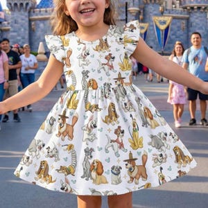 May include: A white dress with a pattern of cartoon dogs, including Lady and Tramp, with ruffle sleeves. The dress is knee-length and has a flared skirt. The background shows a theme park with people.