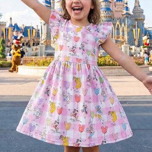 Könnte beinhalten: Ein rosa-weiß kariertes Kleid mit Disney-Figuren und Luftballons. Das Kleid hat kurze Ärmel mit Rüschenkanten und eine taillierte Taille. Zu den Figuren gehören Mickey, Minnie, Donald und Pluto. Im Hintergrund ist ein Schloss zu sehen.