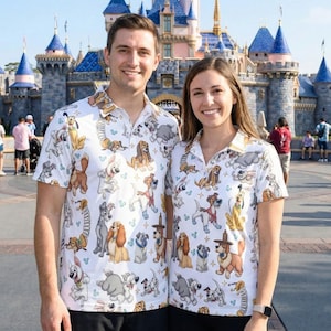 Puede incluir: Camisas blancas a juego con un colorido estampado de perros de dibujos animados. Las camisas presentan varias razas de perros y personajes de Disney. El fondo muestra un castillo, lo que sugiere un parque temático.