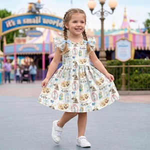 May include: A young girl in a patterned dress with short, ruffled sleeves and blue bows. She wears white trainers and smiles. The dress features cartoon characters. The background shows a theme park with a sign that says "It's a small world".