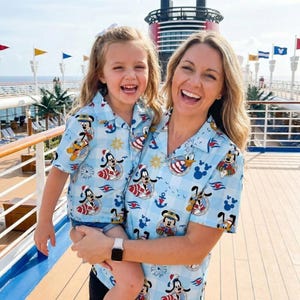 Op de afbeelding: Bijpassende lichtblauwe overhemden met een Disney-personageprint. De shirts hebben een patroon van Mickey Mouse, Donald Duck en Goofy in nautische thema's. De shirts worden gedragen door een vrouw en een kind.