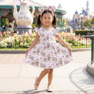 May include: A young girl wearing a white dress with a pink and gold Minnie Mouse teacup pattern, matching headband, and pink shoes. The dress has flutter sleeves. The background includes a Mad Tea Party ride and a castle.