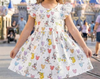 May include: A white dress with a colorful Mickey Mouse and friends balloon print. The dress has short, ruffled sleeves and a flared skirt. The background shows a theme park setting with blurred figures and buildings.