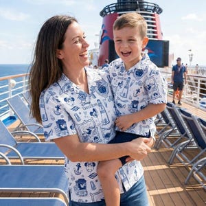 Op de afbeelding: Witte overhemden met een blauw Disney Cruise Line patroon. De shirts hebben nautische elementen, Mickey Mouse en het Disney Cruise Line logo. Een vrouw en een kind dragen de shirts op het dek van een cruiseschip.