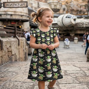 May include: A young girl smiles, wearing a black dress with a repeating pattern of a green alien character in a white bowl. The dress has short, ruffled sleeves. The background includes a Star Wars-themed setting with a spaceship and droids.