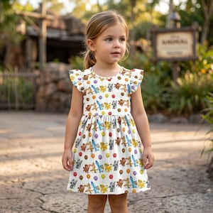May include: A white dress with a pattern of cartoon characters and balloons. The dress has a round neckline and ruffled sleeves. The girl is standing on a paved path in front of a wooden sign.