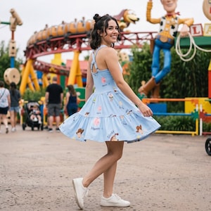 May include: A light blue sleeveless dress with a flared skirt and a crisscross back, featuring an all-over print of Toy Story characters including Woody, Hamm, and Slinky Dog, paired with simple white sneakers for a casual look.
