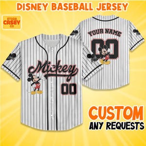 Op de afbeelding: Wit honkbalshirt met zwarte krijtstrepen, met "Mickey" in script en het nummer "00". Het shirt heeft een Mickey Mouse-afbeelding en de tekst "Disney Baseball Jersey". De achterkant heeft ruimte voor een naam en nummer.
