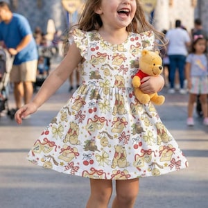 Puede incluir: Un vestido blanco con un estampado de Winnie the Pooh, con el personaje, lazos y otros elementos temáticos. El vestido tiene mangas cortas y una falda acampanada. Un niño sostiene un peluche de Winnie the Pooh.
