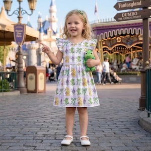May include: A young girl wearing a lavender dress with a repeating pattern of a princess, a frog, and bows. The dress has ruffled sleeves and is paired with white shoes. She holds a green frog plush toy and points towards a sign for Fantasyland.