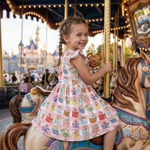 Puede incluir: Una niña sonríe montada en un caballo de carrusel, con un vestido rosa con estampado de libro de cuentos. El carrusel tiene postes dorados y luces. Sostiene un pretzel con forma de Mickey Mouse. Al fondo, un castillo.