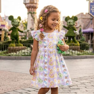 May include: A young girl wearing a lavender and white checkered dress with a Rapunzel print, holding a green plush frog. The dress has flutter sleeves and is adorned with floral hair accessories. The background features topiary figures.