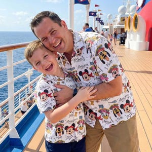 Op de afbeelding: Een vader en kind omarmen elkaar, beiden dragen bijpassende shirts met korte mouwen. De shirts zijn wit met een herhaald patroon van Disney-personages, waaronder Mickey Mouse, Minnie Mouse en Pluto. De achtergrond toont een dek van een cruiseschip en de zee.