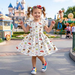 Könnte beinhalten: Ein junges Mädchen trägt ein weißes Kleid mit einem bunten Toy Story-Charakterdruck. Das Kleid hat flatternde Ärmel und einen ausgestellten Rock. Sie trägt bunte Turnschuhe und ein rotes Haaraccessoire. Im Hintergrund ein Vergnügungspark.