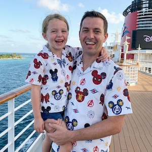 Op de afbeelding: Een vader en kind dragen bijpassende witte overhemden met een patroon van Mickey Mouse, Spider-Man en Avengers-afbeeldingen. De achtergrond toont een dek van een cruiseschip en de zee.