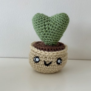 May include: A crocheted heart-shaped plant in a brown and beige pot with a smiling face. The plant is green and the pot has black eyes.