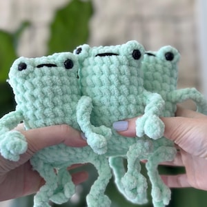 Op de afbeelding: Drie gehaakte groene kikker knuffels met zwarte ogen. De kikkers zitten op elkaar.