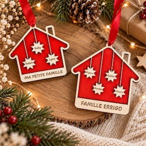 May include: Two house-shaped wooden ornaments with red centers and natural wood borders. Each hangs from a red ribbon. The left ornament features three star-shaped cutouts labeled Sally, Salem, and Lysa, with the text MA PETITE FAMILLE. The right ornament has four star-shaped cutouts labeled Pietro, Dalila, Martika, and Azzura, with the text FAMILLE ERRIGO.