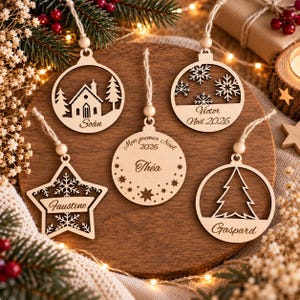 May include: Five light-colored wooden Christmas ornaments with twine hangers. Designs include a house with trees, snowflakes with the text Victor Noël 2026, a star with the name Faustine, a circle with Mon premier Noël 2026 and Théa, and a tree with the name Gaspard.