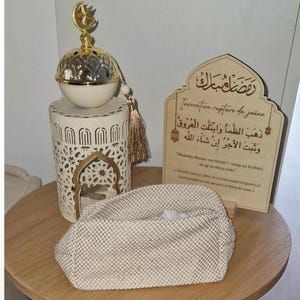 May include: A cream-colored textured pouch sits before a white metal lantern topped with a gold crescent moon and star ornament. Beside it stands a wooden plaque featuring Arabic calligraphy and French text regarding the Ramadan fast breaking.