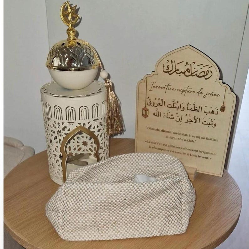 May include: A cream-colored textured pouch sits before a white metal lantern topped with a gold crescent moon and star ornament. Beside it stands a wooden plaque featuring Arabic calligraphy and French text regarding the Ramadan fast breaking.