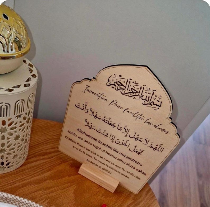 May include: A light-colored wooden plaque with an arched top, featuring engraved Arabic calligraphy and French text. The inscription includes the phrase Invocation Pour faciliter les choses, followed by Arabic script and phonetic transliteration.