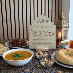 May include: A light-colored wooden sign with an arched top featuring Arabic calligraphy and French text regarding the breaking of the fast. The sign is displayed on a table with a bowl of soup, dates, bread, nuts, and a decorative metal lantern.