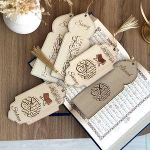 May include: A collection of light-colored wooden bookmarks with arched tops, each featuring engraved Arabic calligraphy, a small red book icon, and a personalized name such as Samy, Ahmed, or Salem, finished with a thin gold-colored tassel.