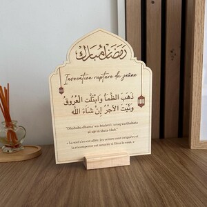 May include: A light-colored wooden arch-shaped sign featuring dark brown printed Arabic calligraphy, the text Invocation rupture du je&ucirc;ne, and the transliterated and translated prayer. Two small lantern illustrations flank the central text.