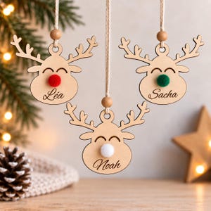 May include: Three light-colored wooden reindeer ornaments hanging from strings. Each reindeer features a fuzzy pom-pom nose in red, green, or white, and is engraved with the names Léa, Sacha, or Noah, along with a small wooden bead on the string.