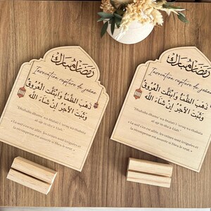 May include: Two identical light-colored wooden plaques with arched tops, each featuring black Arabic calligraphy and French text regarding the breaking of the fast. Each plaque rests on a separate rectangular wooden base stand.