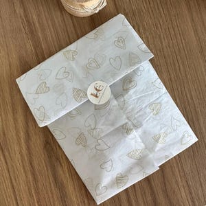 May include: A rectangular package wrapped in white tissue paper featuring a repeating pattern of gold-colored heart outlines. The paper is secured with a small, round white sticker in the center that displays the text "Le Crea" in black script.