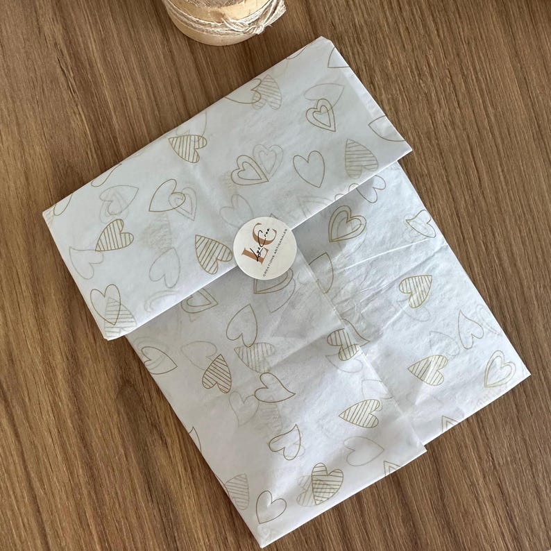 May include: A rectangular package wrapped in white tissue paper featuring a repeating pattern of gold-colored heart outlines. The paper is secured with a small, round white sticker in the center that displays the text "Le Crea" in black script.