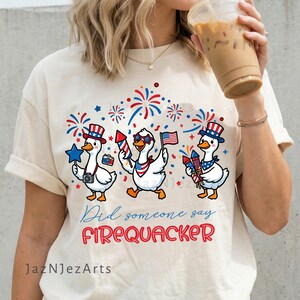 May include: A cream-colored t-shirt featuring three white ducks wearing patriotic hats and accessories, surrounded by red and blue fireworks. The text reads, "Did someone say FIREQUACKER" in blue and red script and block lettering.