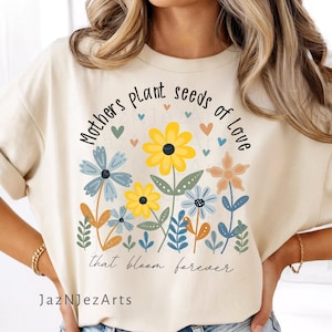Puede incluir: Camiseta color crema con un diseño floral y el texto "Mothers plant seeds of Love that bloom forever." El diseño incluye flores amarillas, azules y naranjas con hojas verdes y pequeños corazones. El nombre del artista, JazNJeZArts, está en la parte inferior.