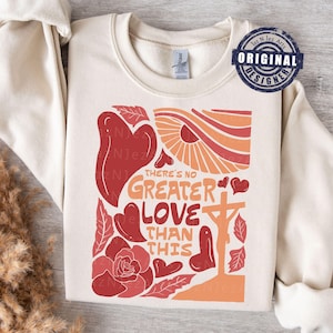 Puede incluir: Una sudadera de color crema con un diseño gráfico rojo y naranja. El gráfico presenta una cruz, corazones, flores y el texto "No hay amor más grande que este".